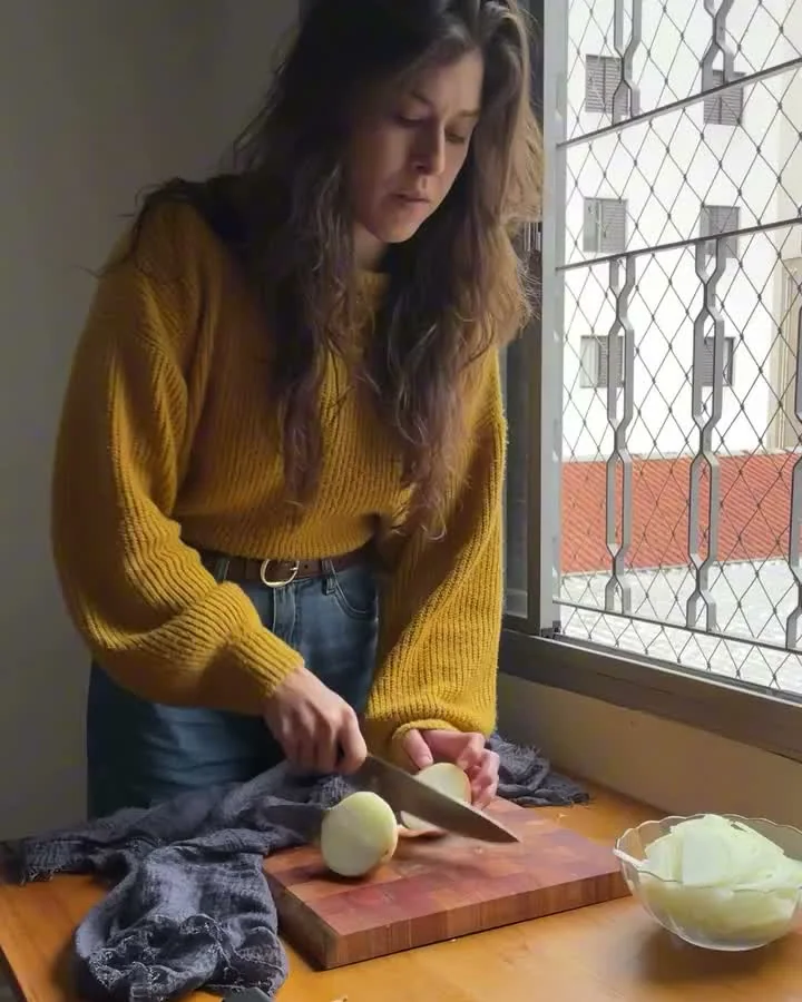 Mica de suéter amarelo cortando uma cebola diante da janela da cozinha